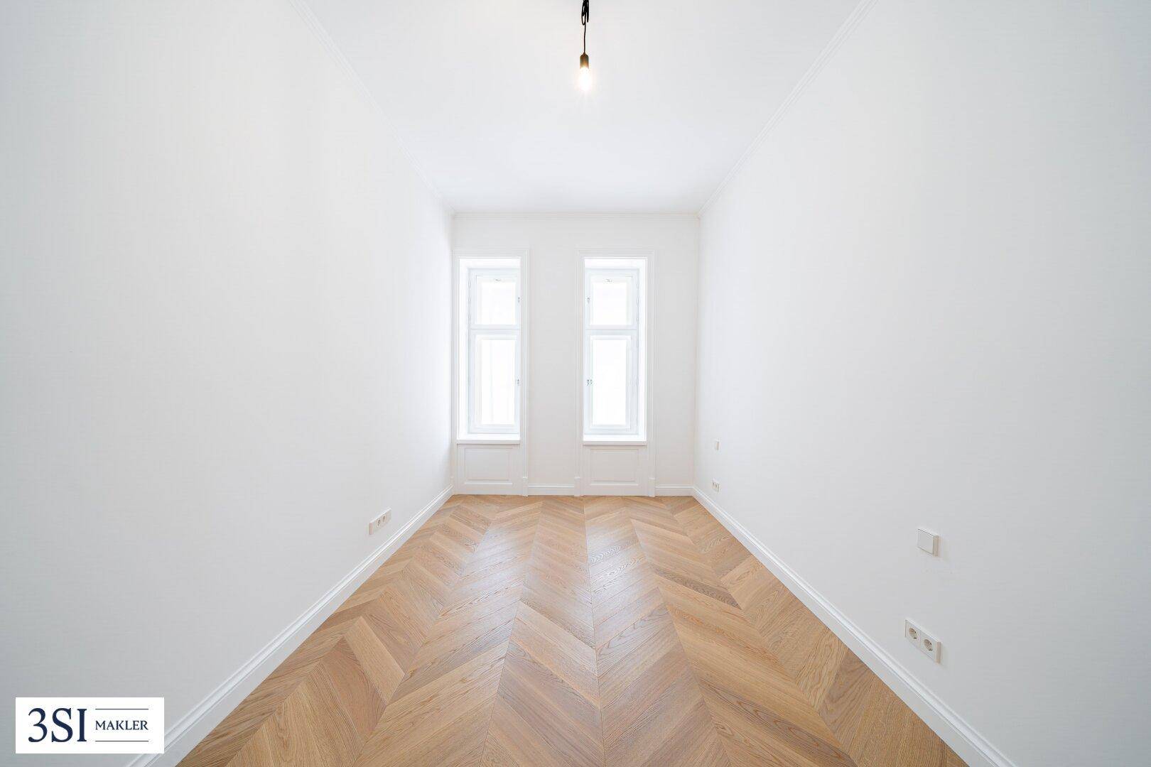 Schmaler, heller Raum mit Fischgrätparkett und zwei Fenstern, ideal als Schlaf- oder Arbeitszimmer.