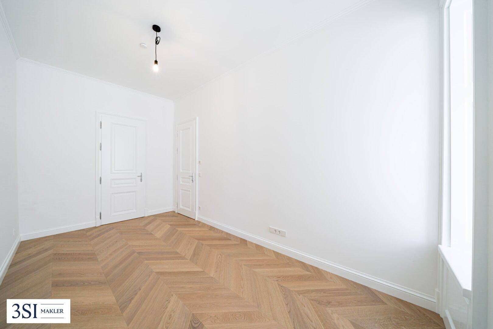 Lichtdurchfluteter Raum mit klassischem Fischgrätparkett und hohen weißen Wänden, ideal für ein Wohnzimmer.