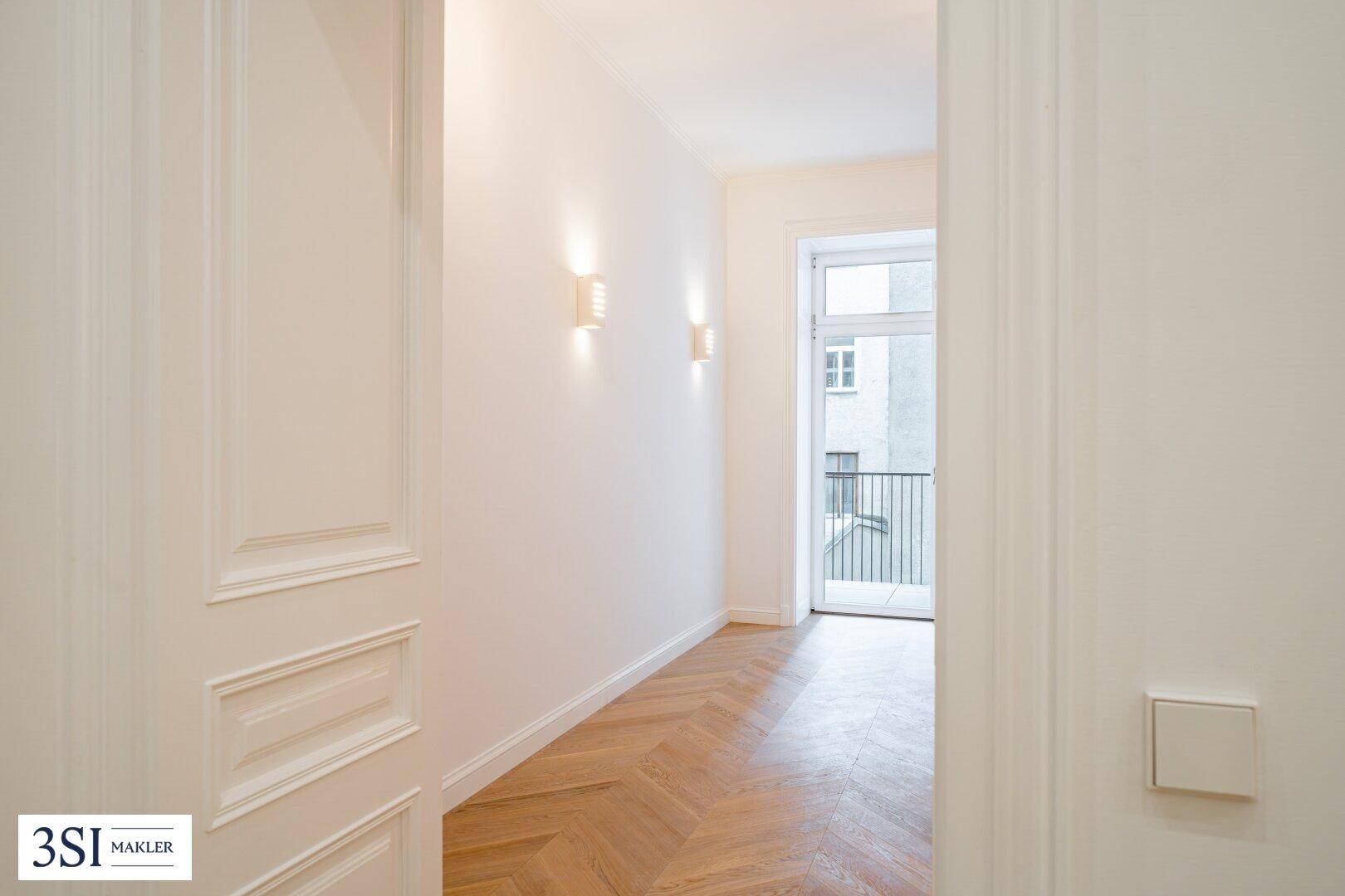 Heller Flur mit Fischgrätparkett und Zugang zu einem Balkon, der viel Licht hereinlässt.