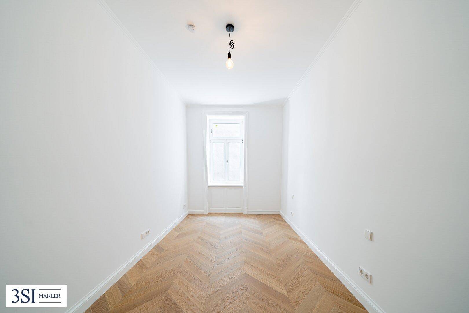 Kleines, helles Zimmer mit Fischgrätparkett, hohen Decken und einem Fenster.