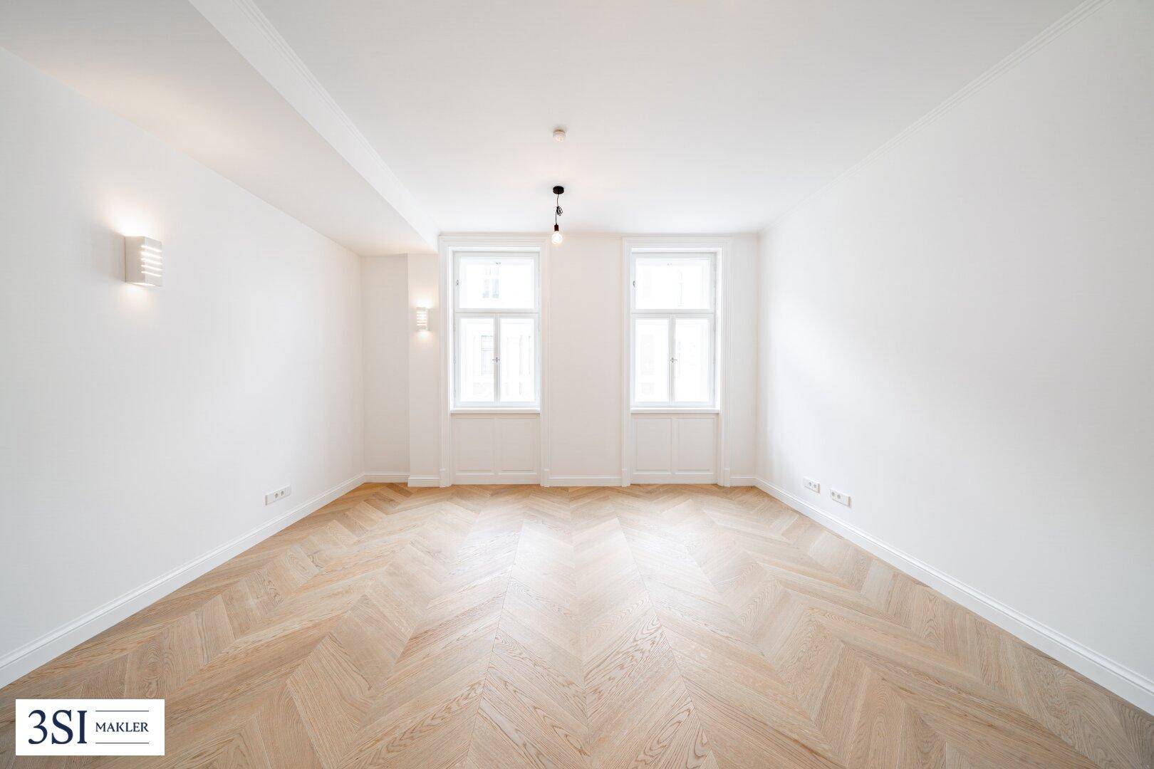 Helles Zimmer mit Fischgrätparkett, hohen Decken und zwei großen Fenstern.