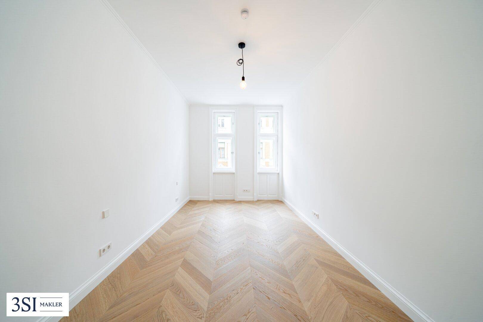 Leeres Zimmer mit Fischgrätparkett, hohen Decken und zwei hohen Fenstern.