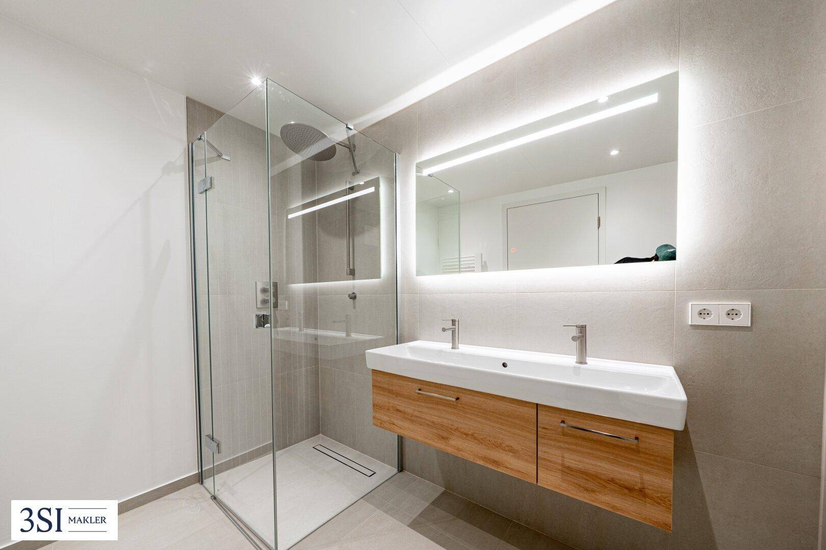 Modernes Badezimmer mit begehbarer Dusche, Doppelwaschbecken und beleuchtetem Spiegel.