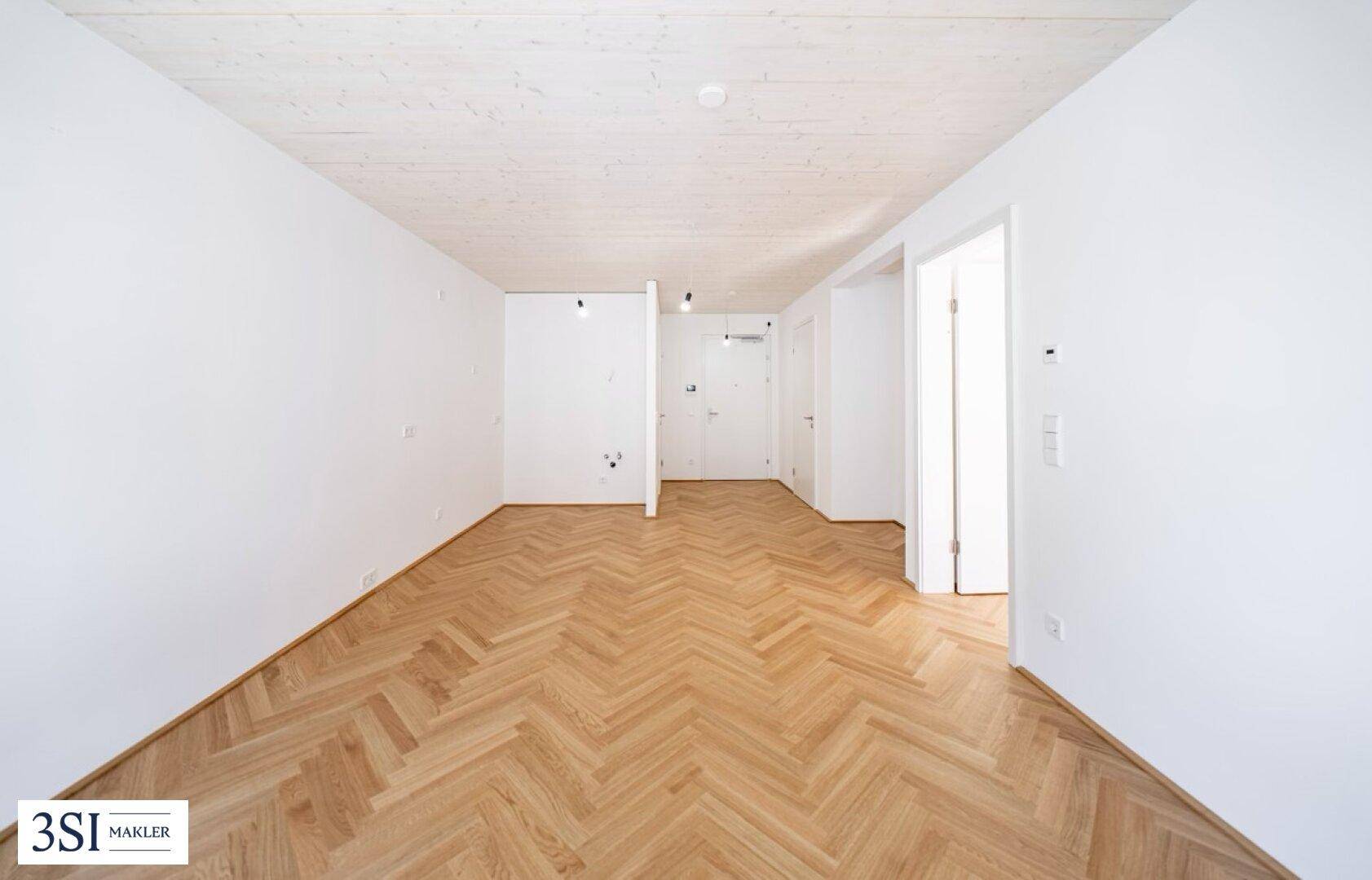 Geräumiger, leerer Raum mit hellem Fischgrätparkett und weiß gestrichenen Wänden.