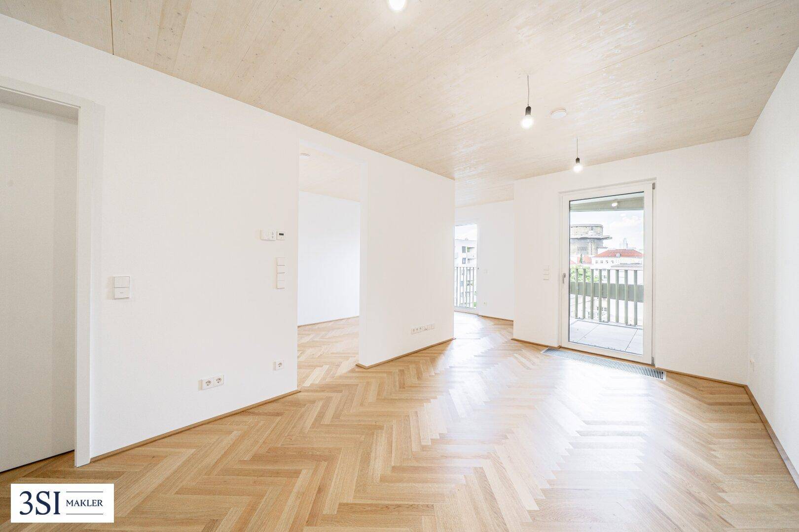 Leerer, lichtdurchfluteter Raum mit Fischgrätparkett und Zugang zu einem Balkon.