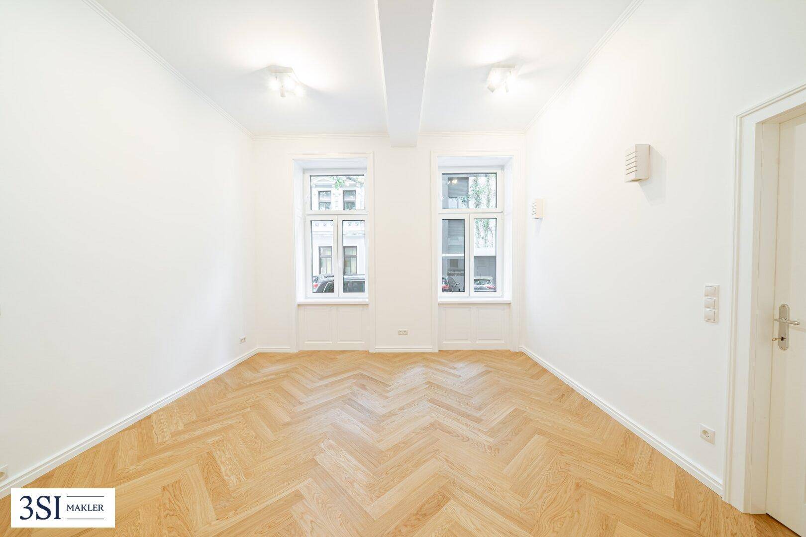 Helles, leeres Schlafzimmer mit Fischgrätparkett und zwei großen Fenstern, ideal für individuelle Gestaltung.