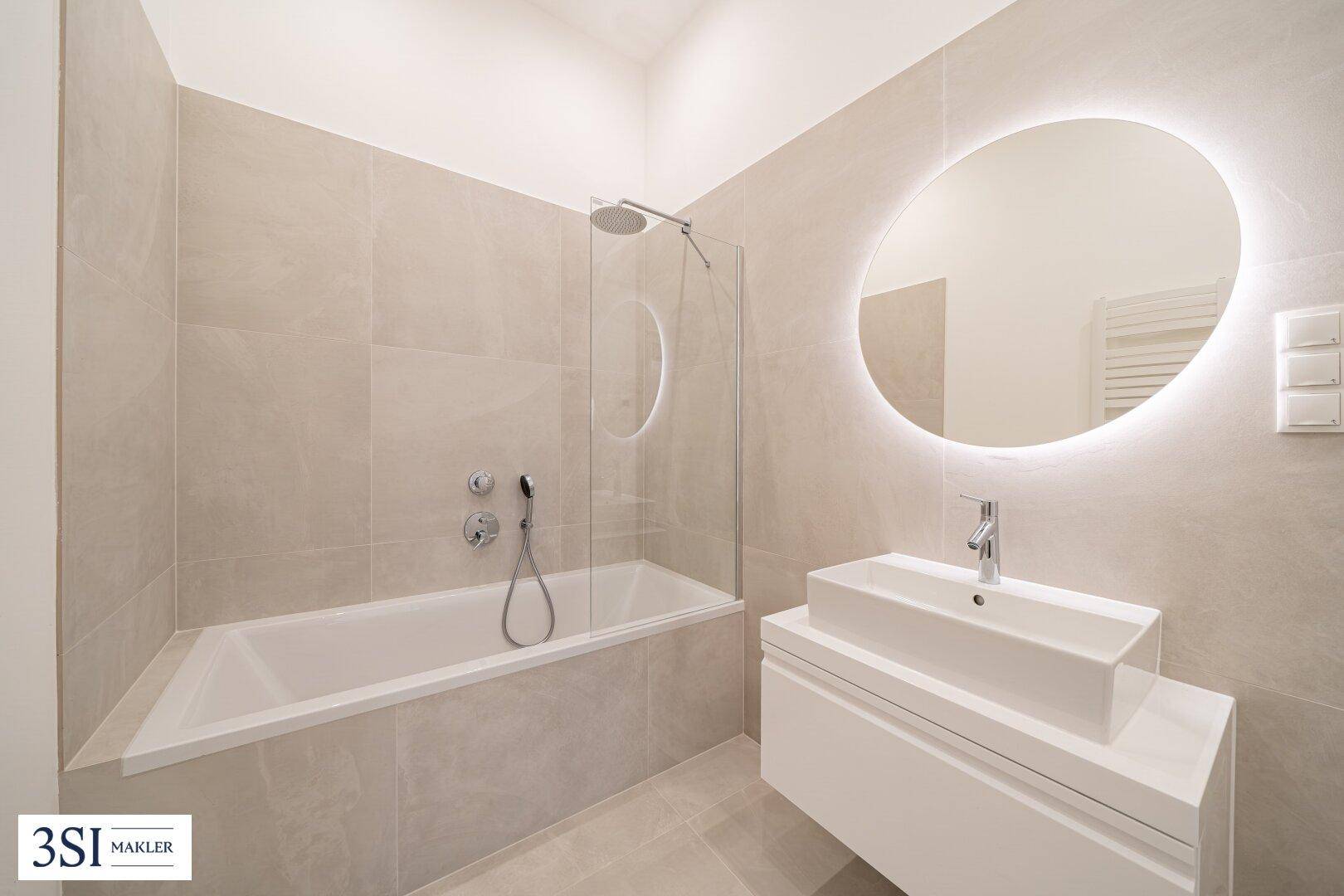 Modernes Badezimmer mit Badewanne, integrierter Dusche, beleuchtetem Spiegel und eleganten beigen Fliesen.