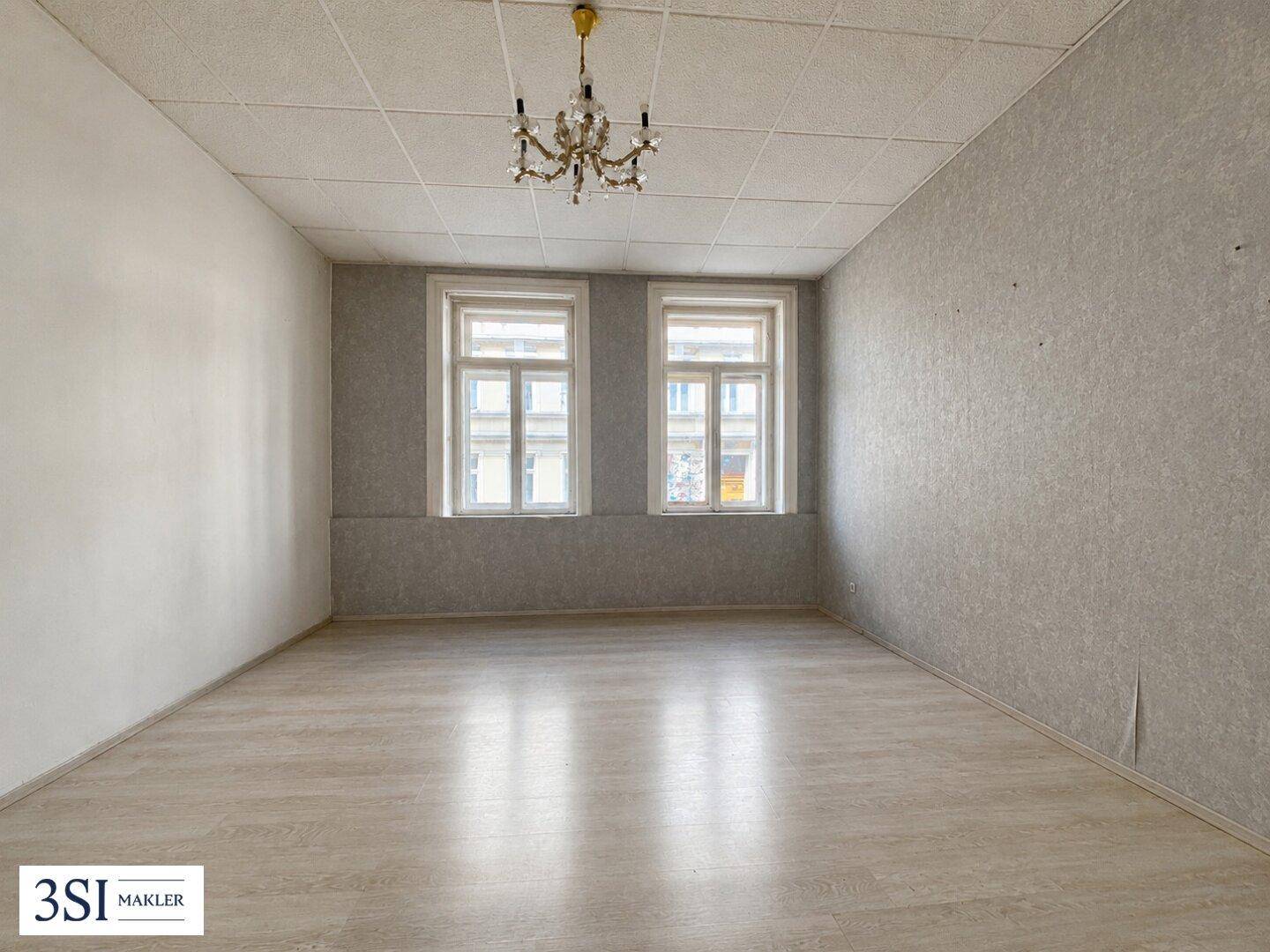 Leeres Wohnzimmer mit hellem Laminatboden, hohen Decken und zwei großen Fenstern.