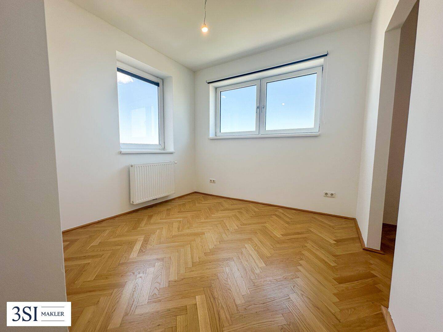 Freundliches Zimmer mit Fischgrätparkett und großen Fenstern, die viel Licht hereinlassen.