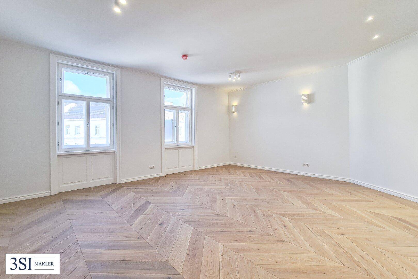 Heller, leerer Raum mit Fischgrätparkett und hohen Fenstern, ideal als Wohnzimmer.
