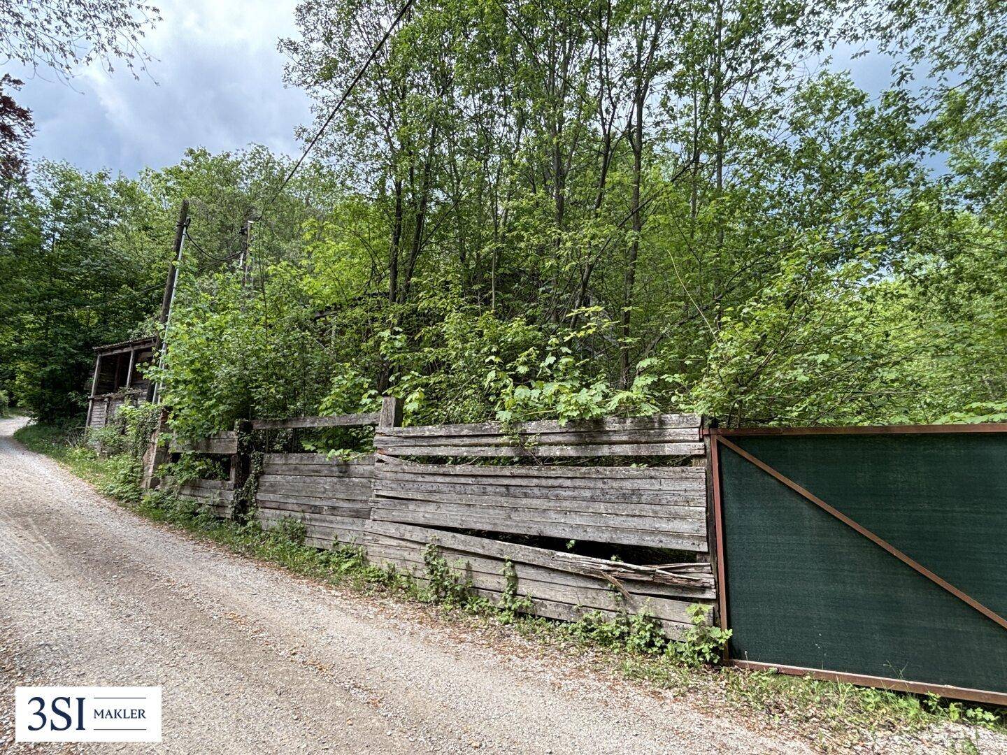 Schotterweg führt entlang eines alten Holzzauns und eines grünen Tores zu einem bewaldeten Grundstück.