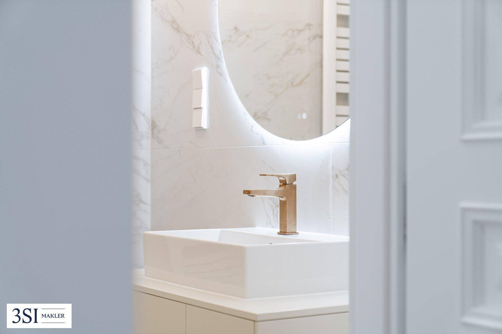 Modernes Badezimmer mit elegantem Waschtisch, goldfarbener Armatur und beleuchtetem Rundspiegel.