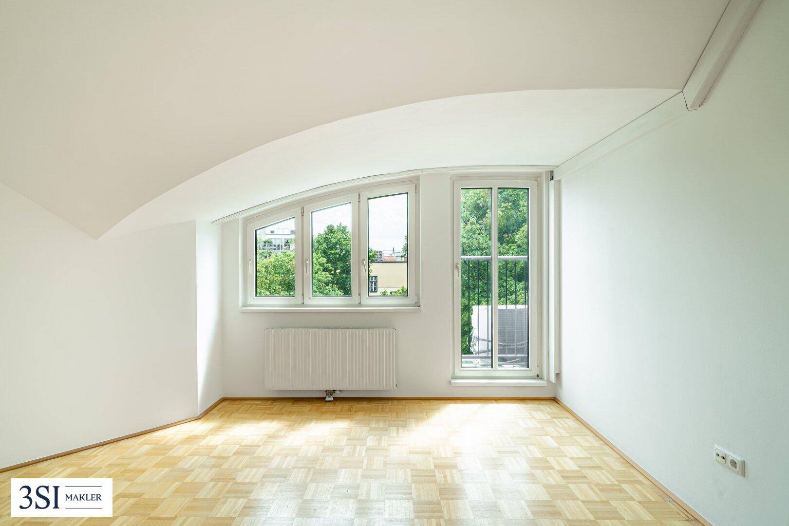 Helles Zimmer mit Parkettboden, Dachschräge und Fenstern, die einen Blick ins Grüne bieten.