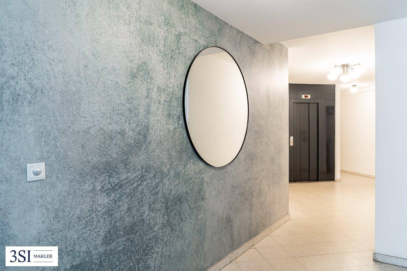 Moderner Flur mit strukturierter Wand, rundem Spiegel und Zugang zu einem Aufzug.