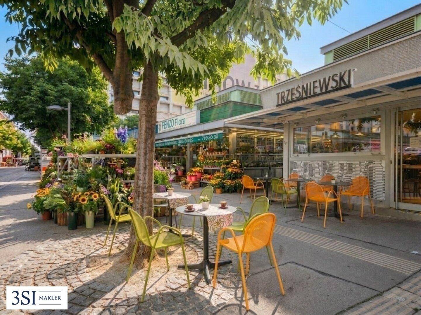 Lebhafte Straßenszene mit Cafétischen, Blumenladen und Geschäften unter grünen Bäumen.