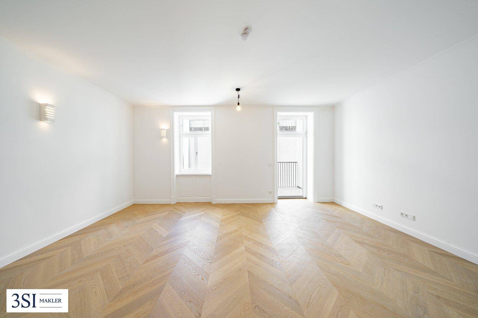 Heller, leerer Raum mit Fischgrätparkett, hohen Decken und Zugang zu einem französischen Balkon.