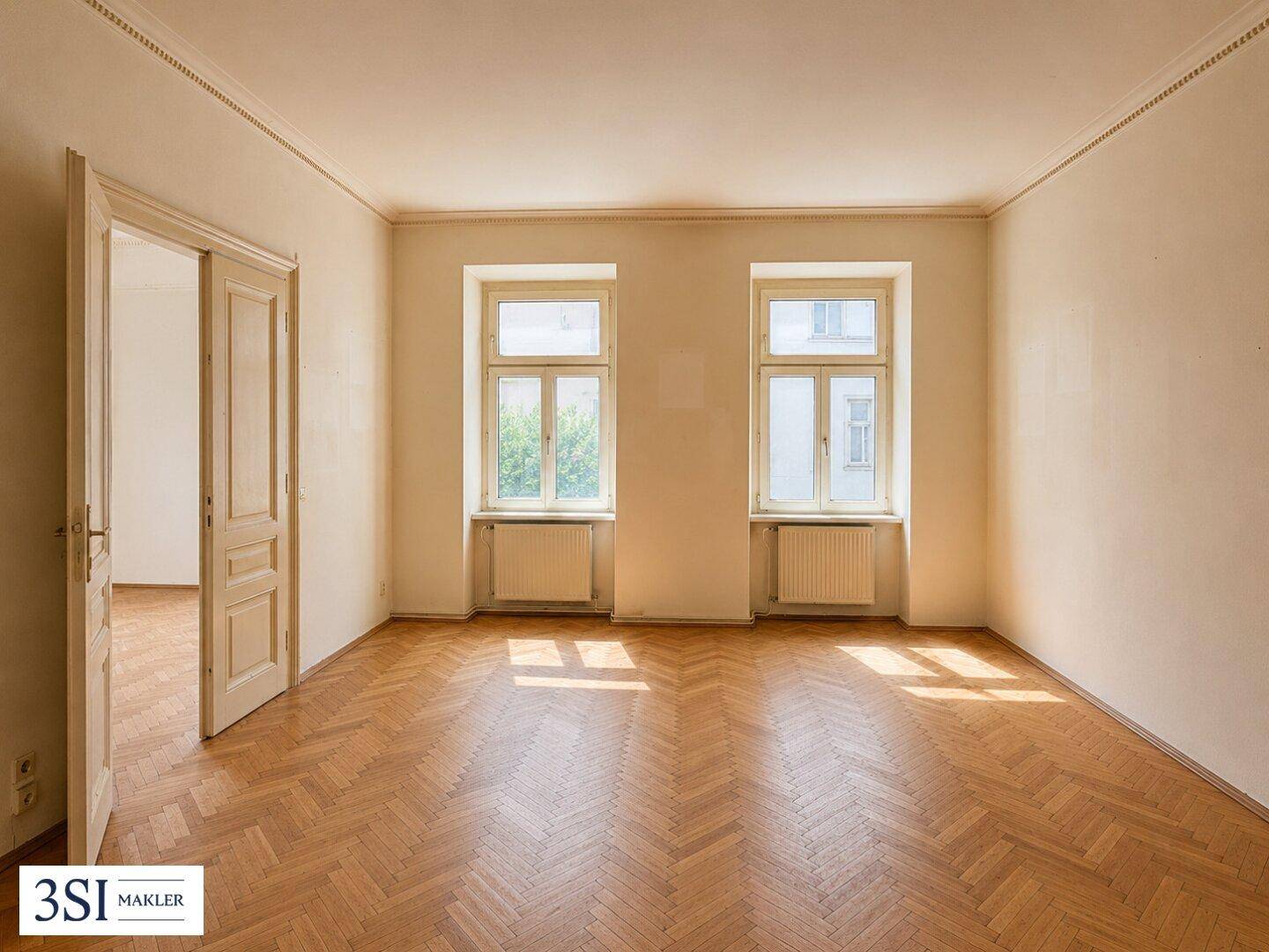 Geräumiges, unmöbliertes Wohnzimmer mit Fischgrätparkett und hohen Decken.