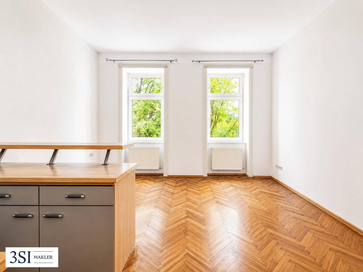 Heller, leerer Raum mit Fischgrätparkett und großen Fenstern mit Blick ins Grüne.