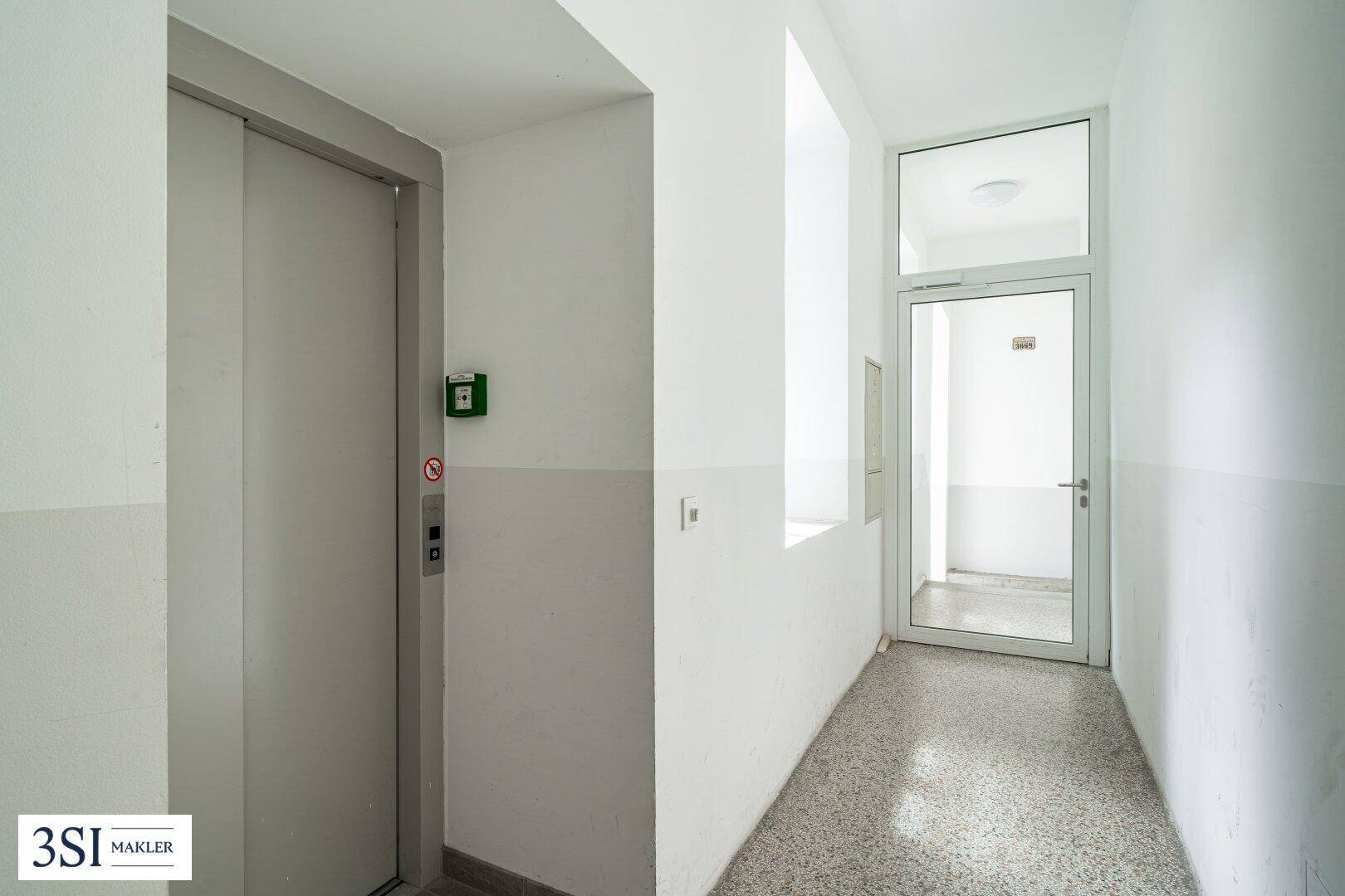 Heller Flur mit Aufzug und einer Glastür, die zu einem weiteren Bereich des Gebäudes führt.