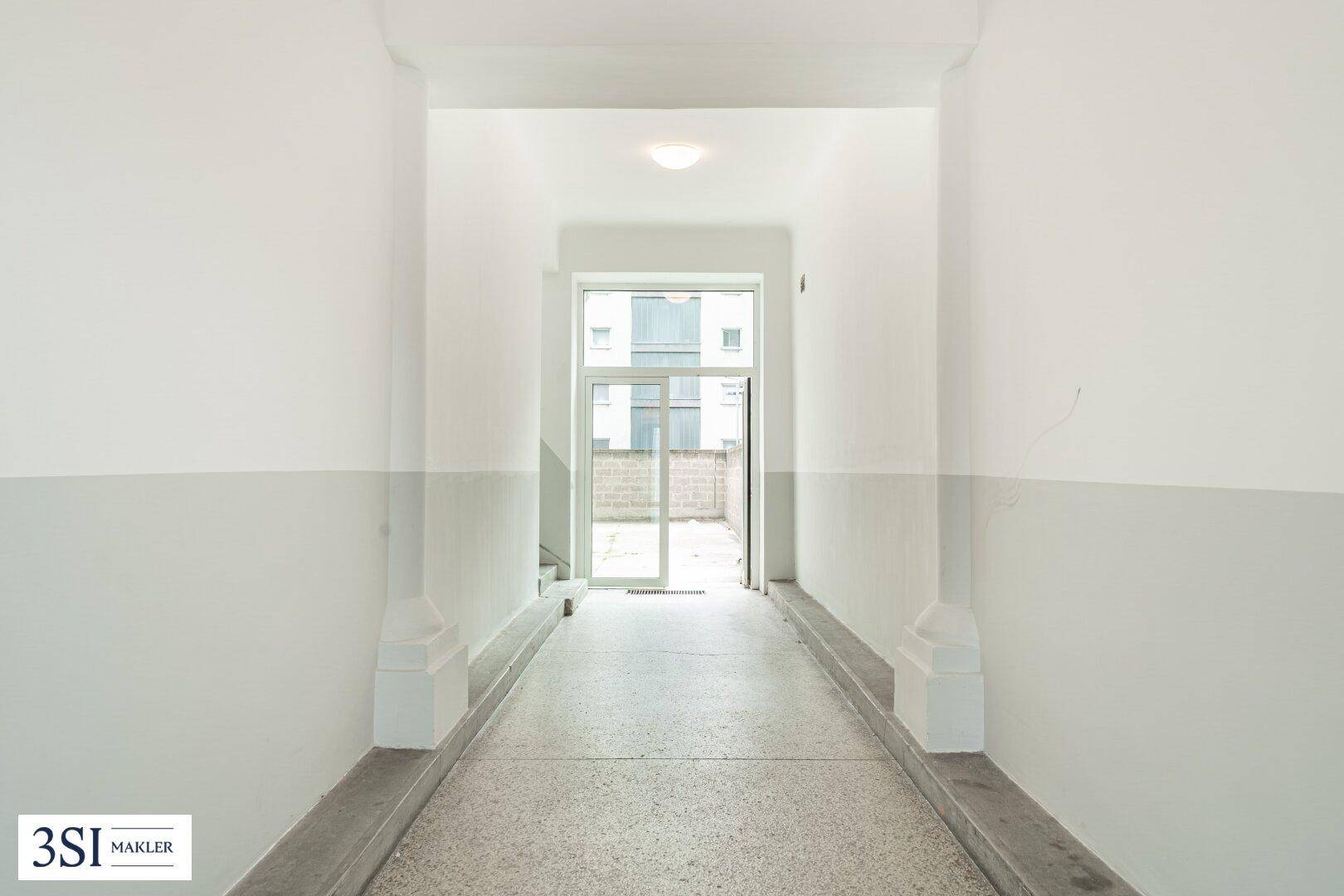 Langer Flur mit Terrazzoboden, der zu einem Ausgang mit großer Glastür und Tageslicht führt.