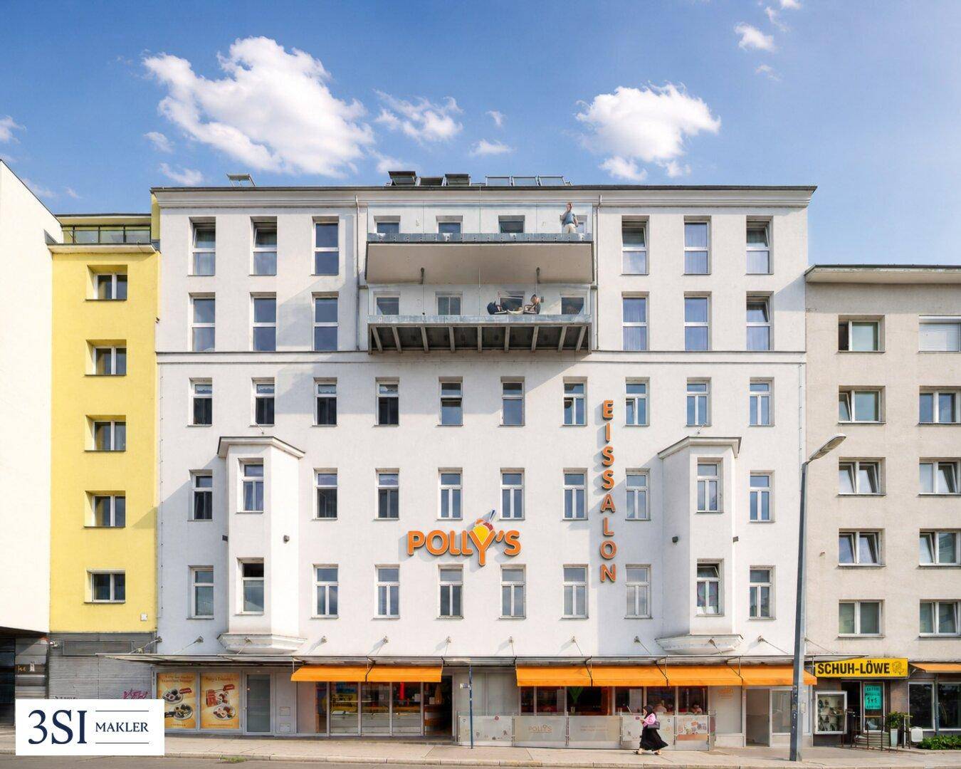 Mehrstöckiges Gebäude mit Geschäften im Erdgeschoss und einem Balkon im oberen Bereich.