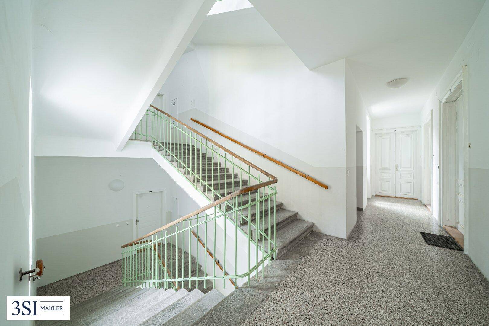 Geräumiges Treppenhaus mit Terrazzoboden, grünem Geländer und viel Tageslicht.