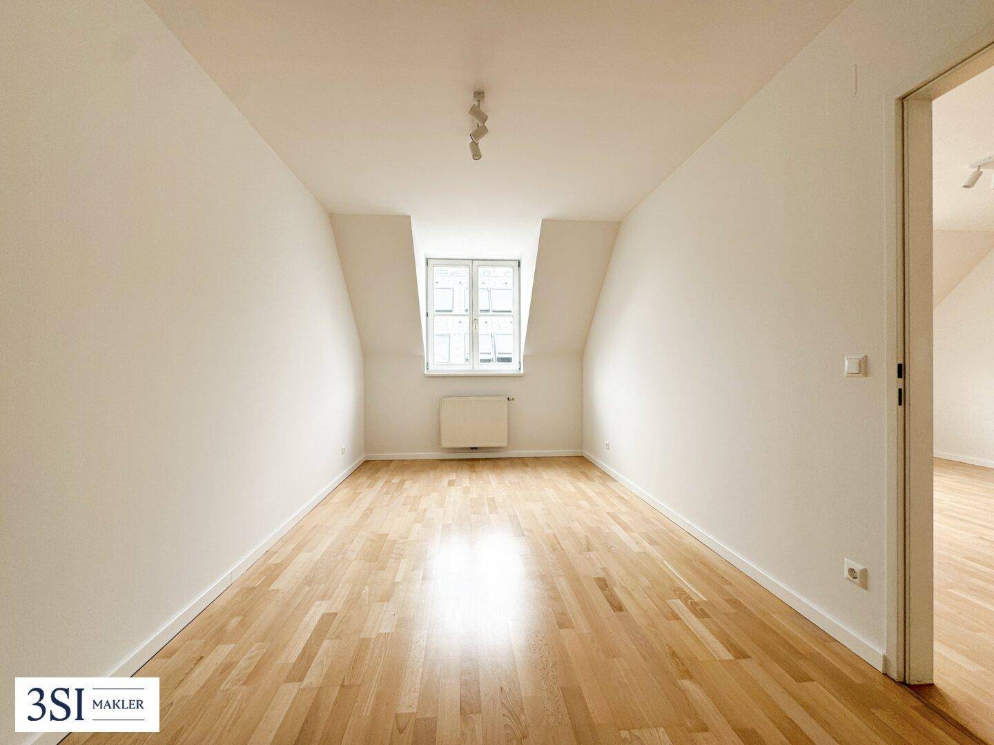 Leeres Zimmer mit Holzboden, einem Fenster und einem Heizkörper, bereit zur individuellen Gestaltung.