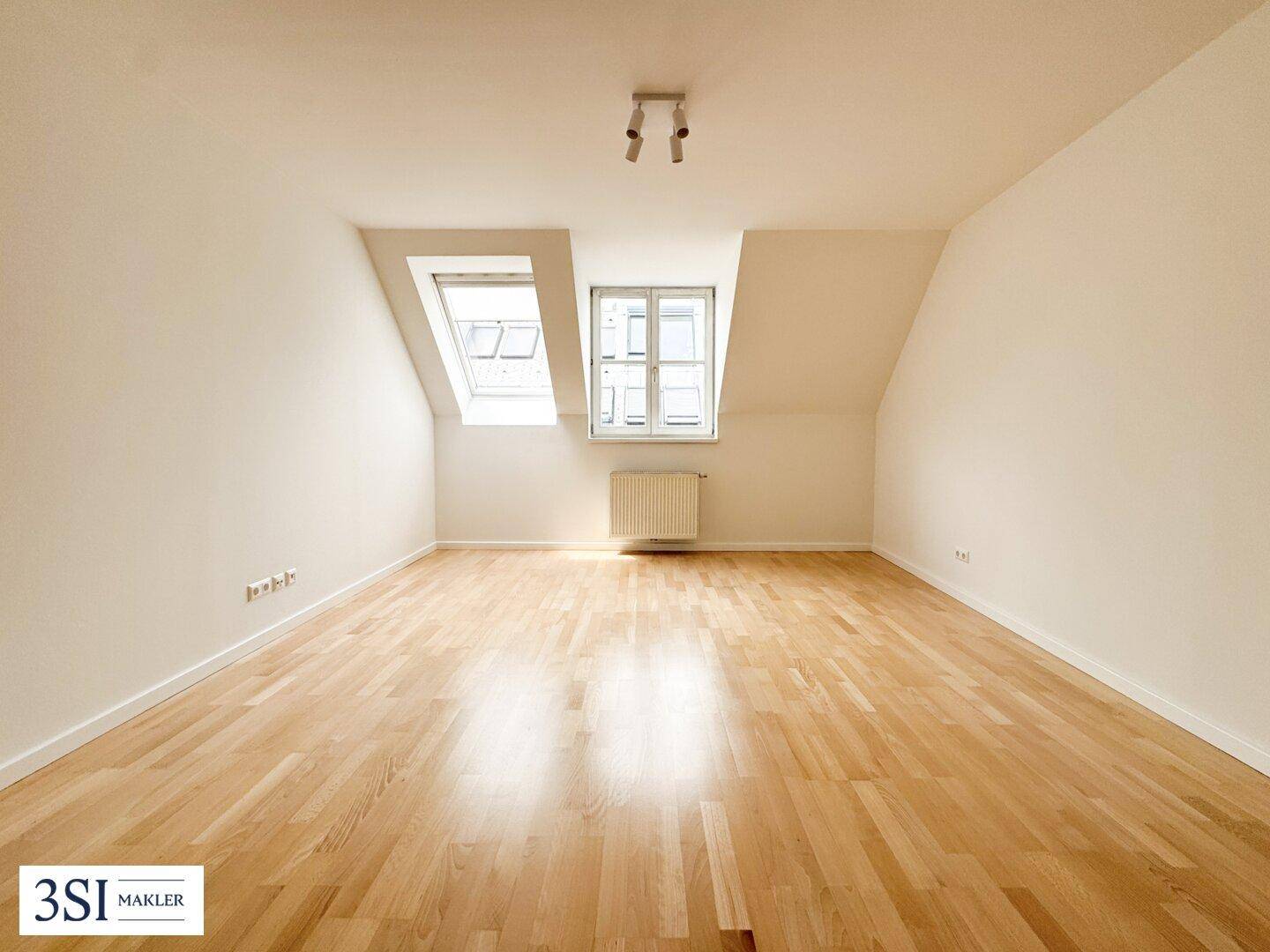 Helles Zimmer im Dachgeschoss mit Holzboden, Dachfenstern und einem Heizkörper unter dem Fenster.