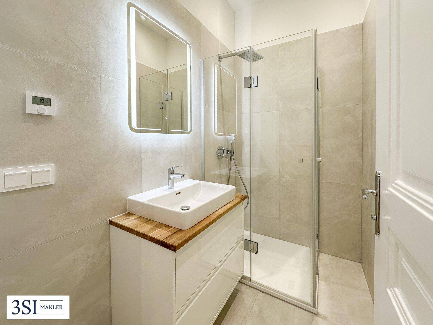 Modernes Badezimmer mit begehbarer Dusche, eleganten Fliesen und beleuchtetem Spiegel.