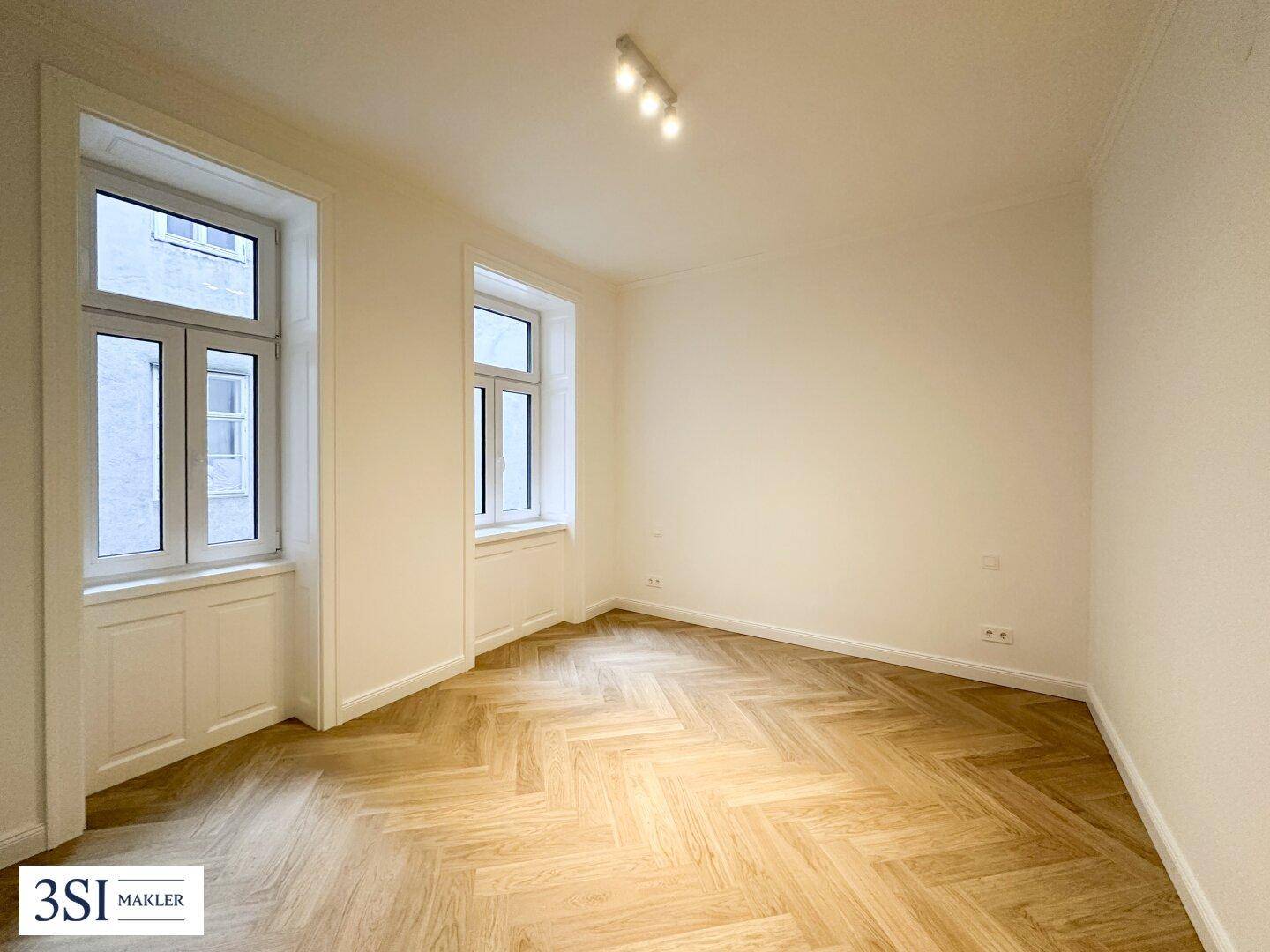 Helles Zimmer mit zwei Fenstern und elegantem Fischgrätparkett, ideal als Schlaf- oder Arbeitszimmer.
