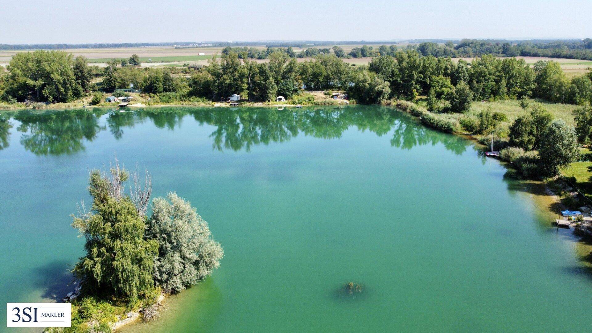 Luftaufnahme eines Sees mit einer kleinen Insel und bewaldeten Ufern in ländlicher Umgebung.