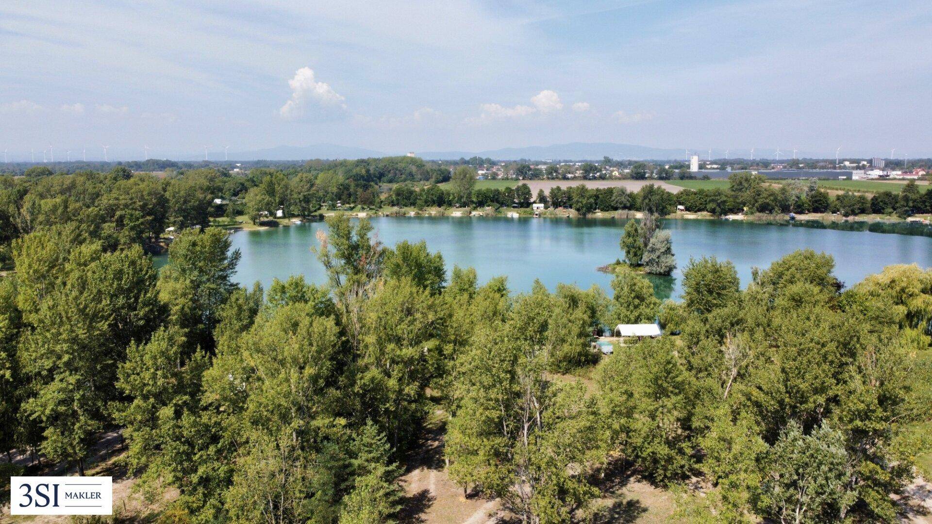 Luftaufnahme eines Sees mit bewaldeten Ufern und einer kleinen Insel, umgeben von grüner Landschaft.