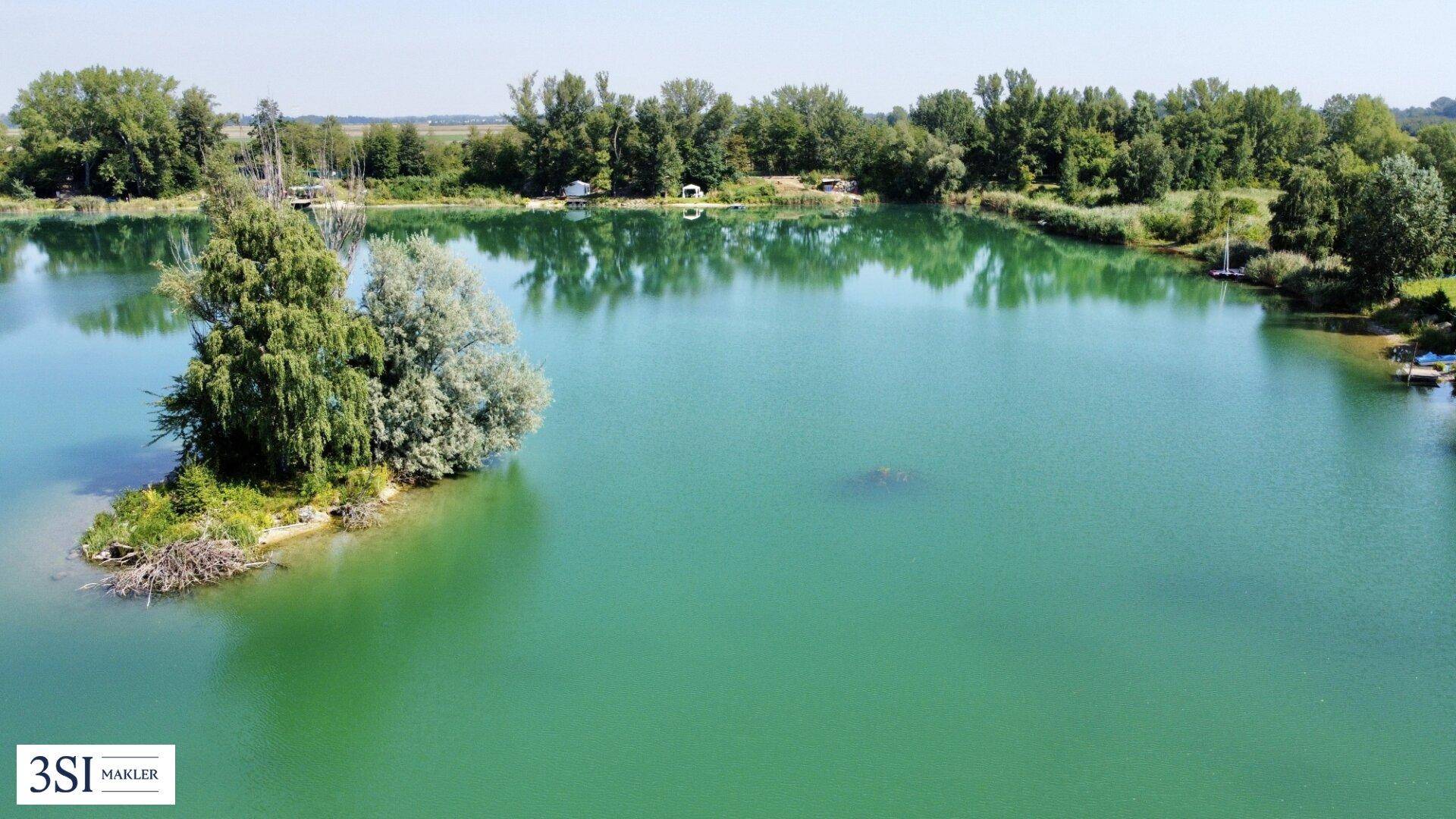 Luftaufnahme eines Sees mit einer kleinen Insel und bewaldeten Ufern in ländlicher Umgebung.