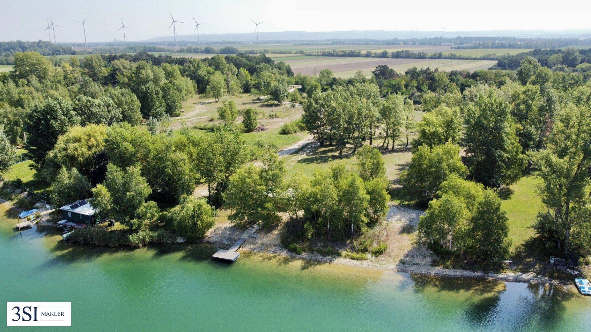 Luftaufnahme eines Sees mit bewaldeten Ufern, kleinen Stegen und Windrädern in der Ferne.