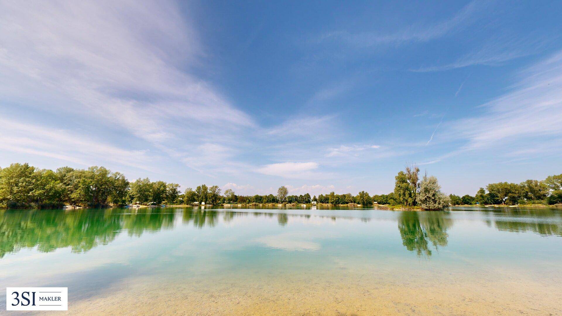 Weitläufiger Blick über einen ruhigen See mit sandigem Ufer und bewaldeter Landschaft unter blauem Himmel.