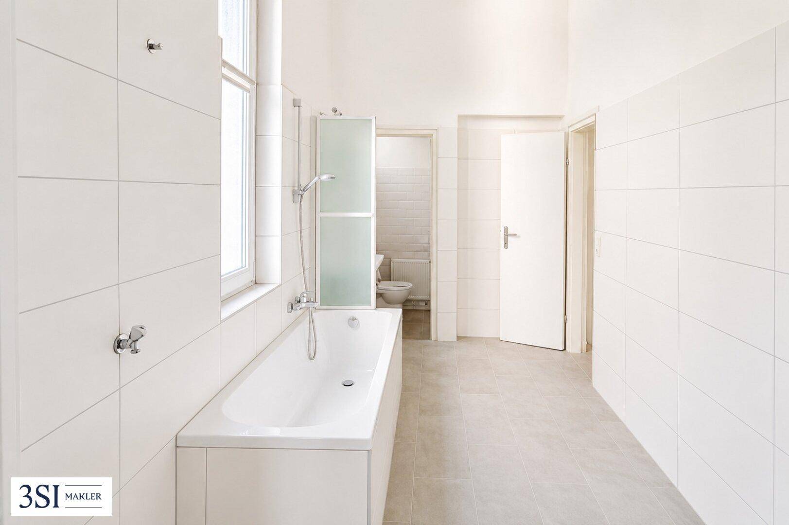 Geräumiges Badezimmer mit weiß gefliesten Wänden, Badewanne und Fenster für Tageslicht.