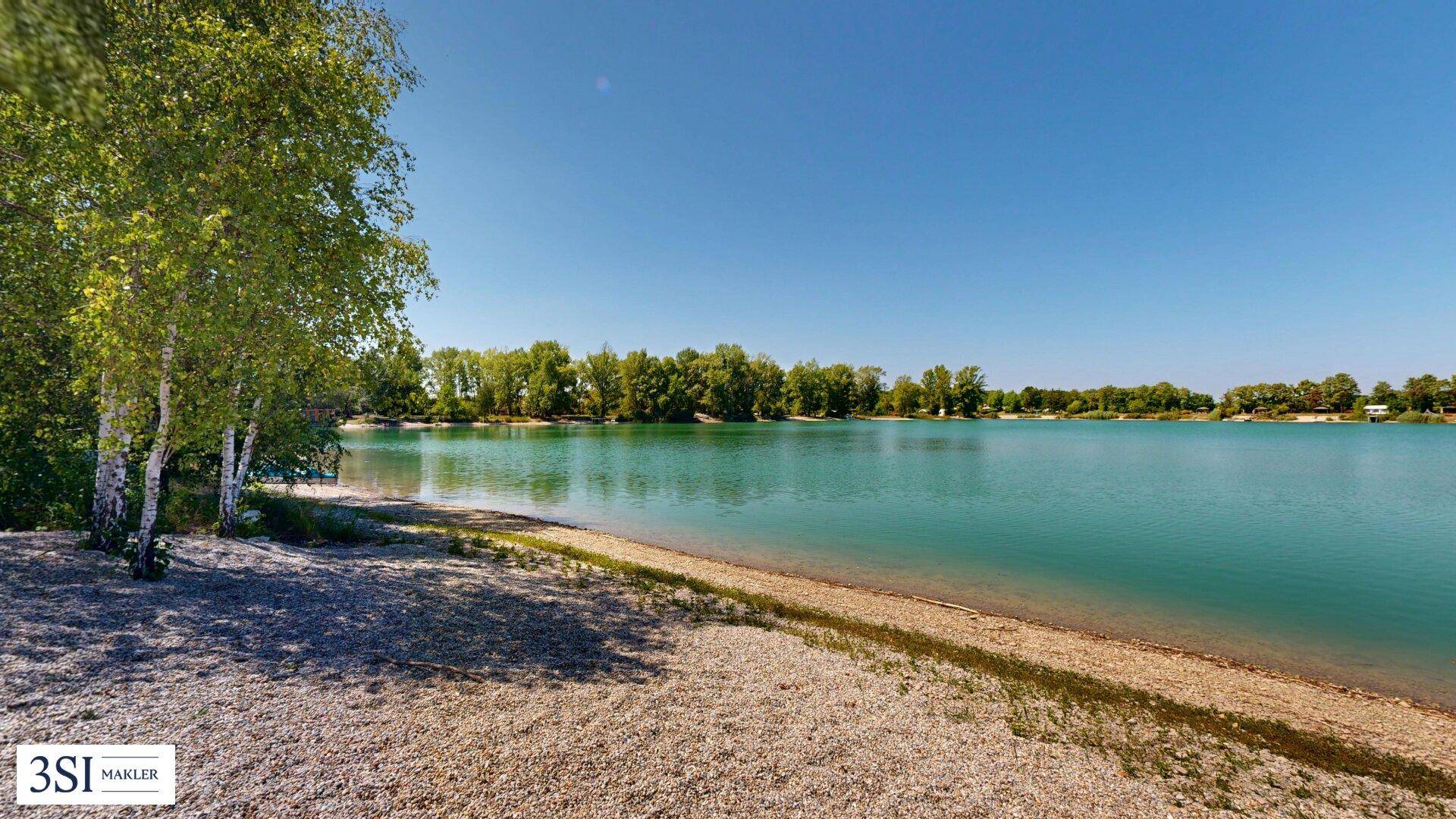 Blick auf den ruhigen See mit klarem Wasser und einem kiesigen Ufer, umgeben von grünen Bäumen.