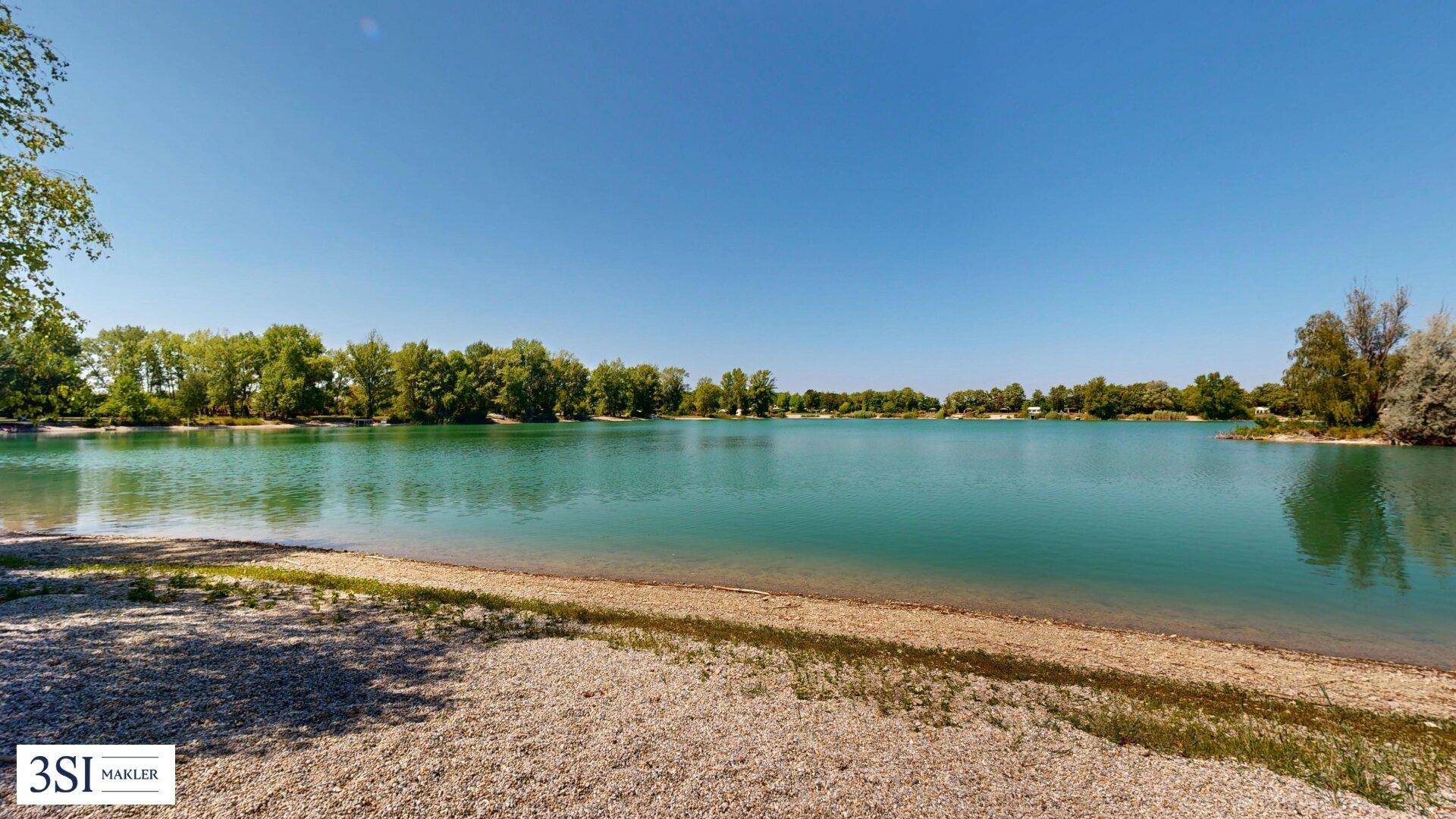 Panoramablick auf einen weitläufigen See mit klarem, türkisfarbenem Wasser und bewaldeten Ufern.