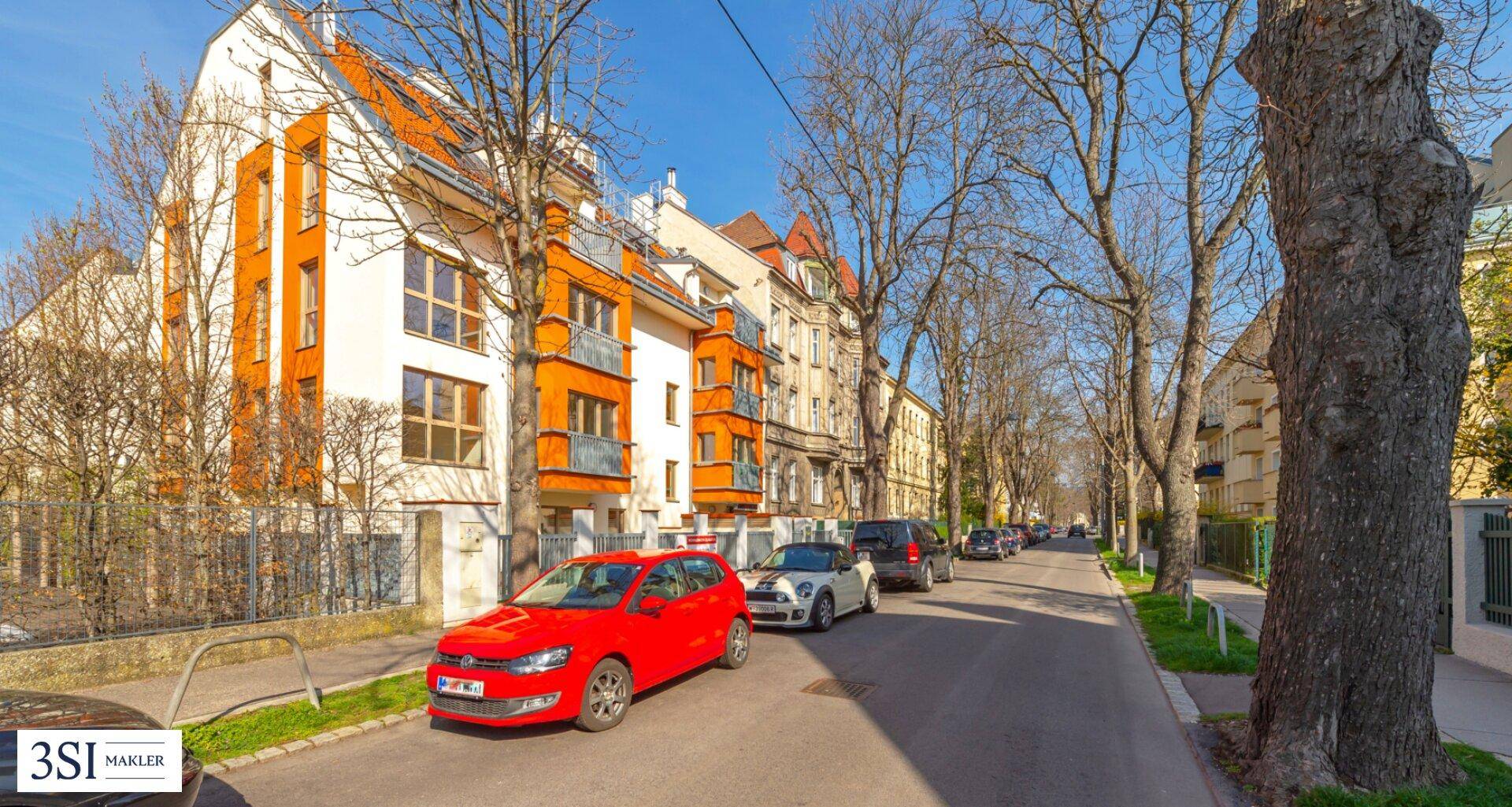Wohnstraße mit geparkten Autos und einem modernen Gebäude mit orangefarbenen Akzenten unter blauem Himmel.