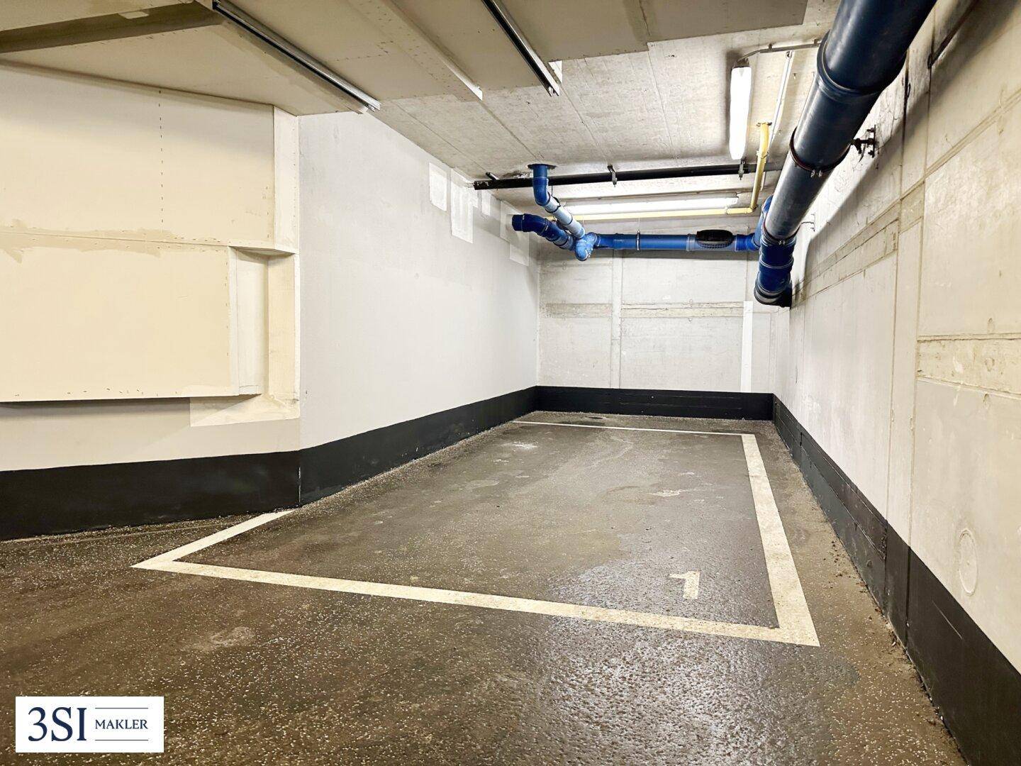 Einzelner Stellplatz in einer Tiefgarage mit blauen Rohren an der Decke und Betonwänden.