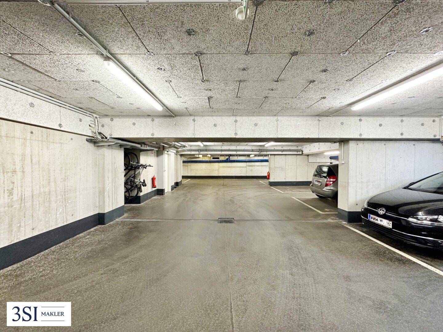 Weitläufige Tiefgarage mit Betonstruktur, Beleuchtung und geparkten Fahrzeugen sowie Fahrradstellplätzen.