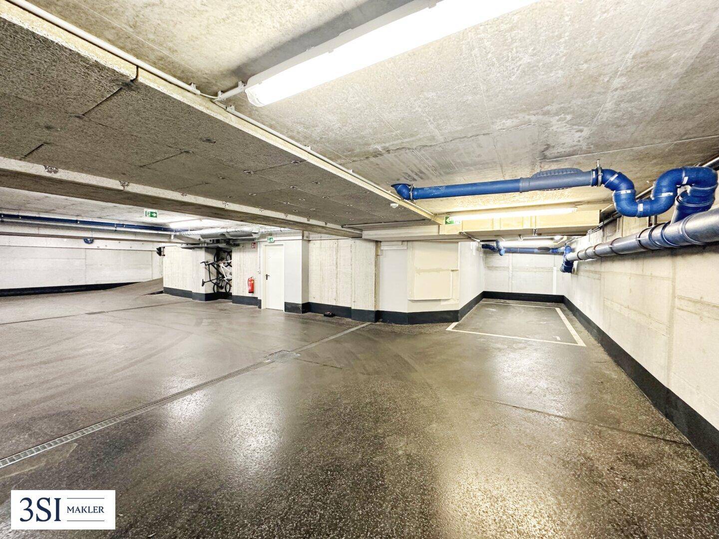 Geräumige Tiefgarage mit Betonwänden, sichtbaren Versorgungsleitungen und Fahrradstellplätzen im Hintergrund.