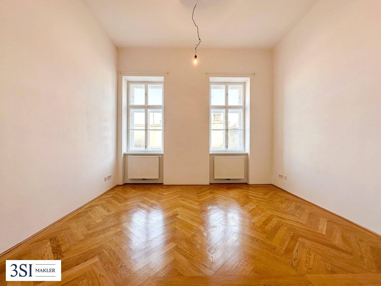 Heller, leerer Wohnraum mit Fischgrätparkett und hohen Fenstern, bereit zur individuellen Gestaltung.