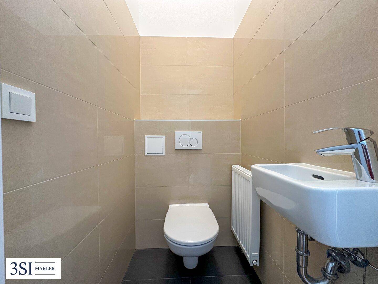 Separates Gäste-WC mit Wand-WC, kleinem Waschbecken und beigefarbenen Fliesen.
