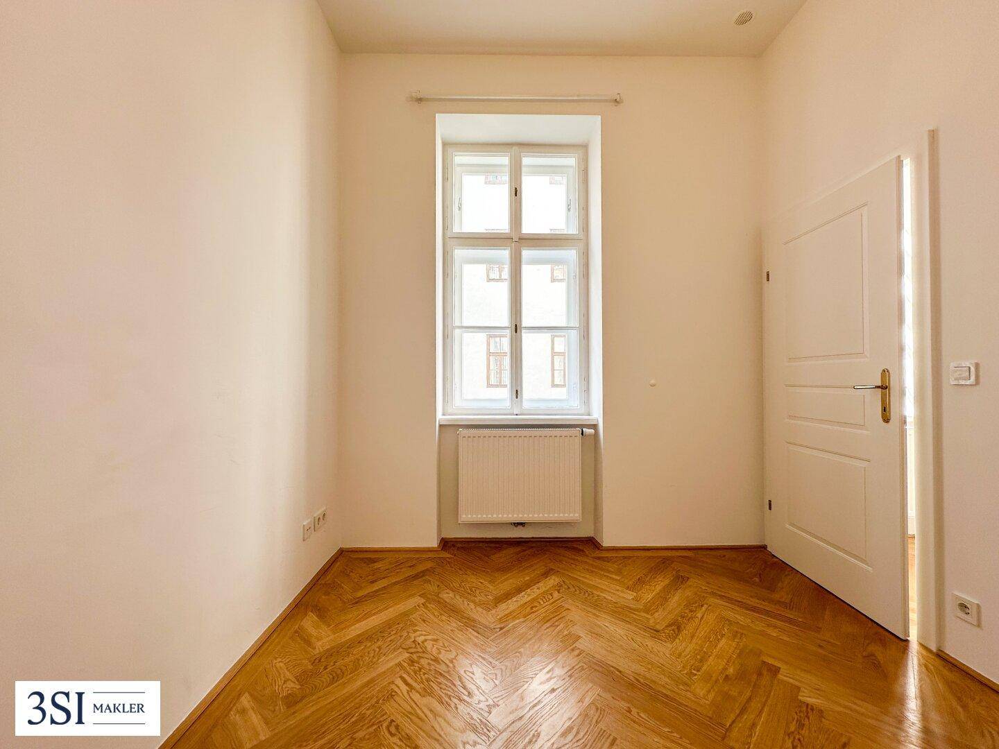 Leeres Schlafzimmer mit Fischgrätparkett und einem großen Fenster, ideal für individuelle Einrichtung.