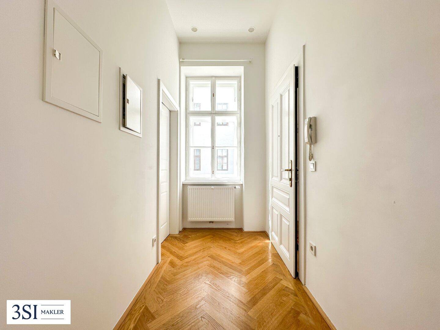 Heller Flur mit Fischgrätparkett und einem Fenster, das Tageslicht hereinlässt.