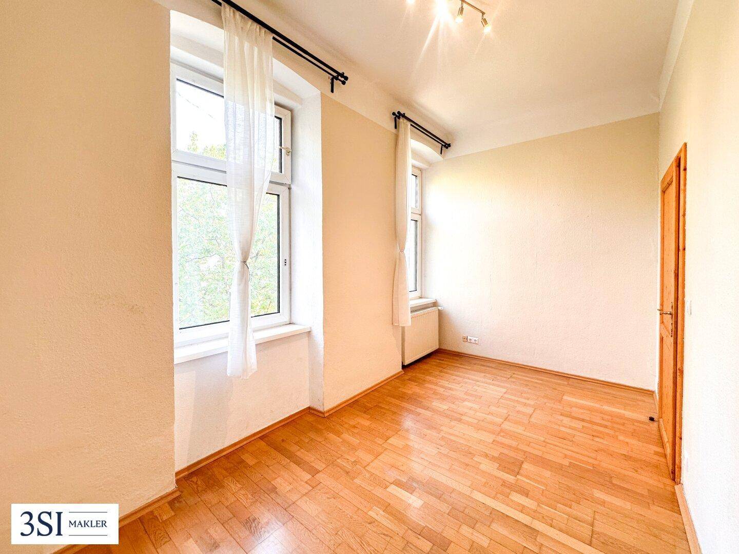 Geräumiges Wohnzimmer mit Holzboden und zwei Fenstern, die den Raum gut belichten.
