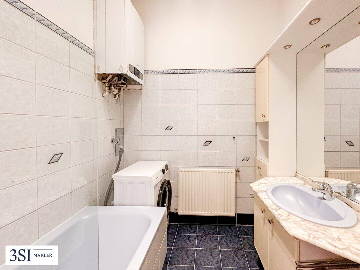 Badezimmer mit Badewanne, Waschbecken und Waschmaschine, gefliest in hellen und dunklen Tönen.
