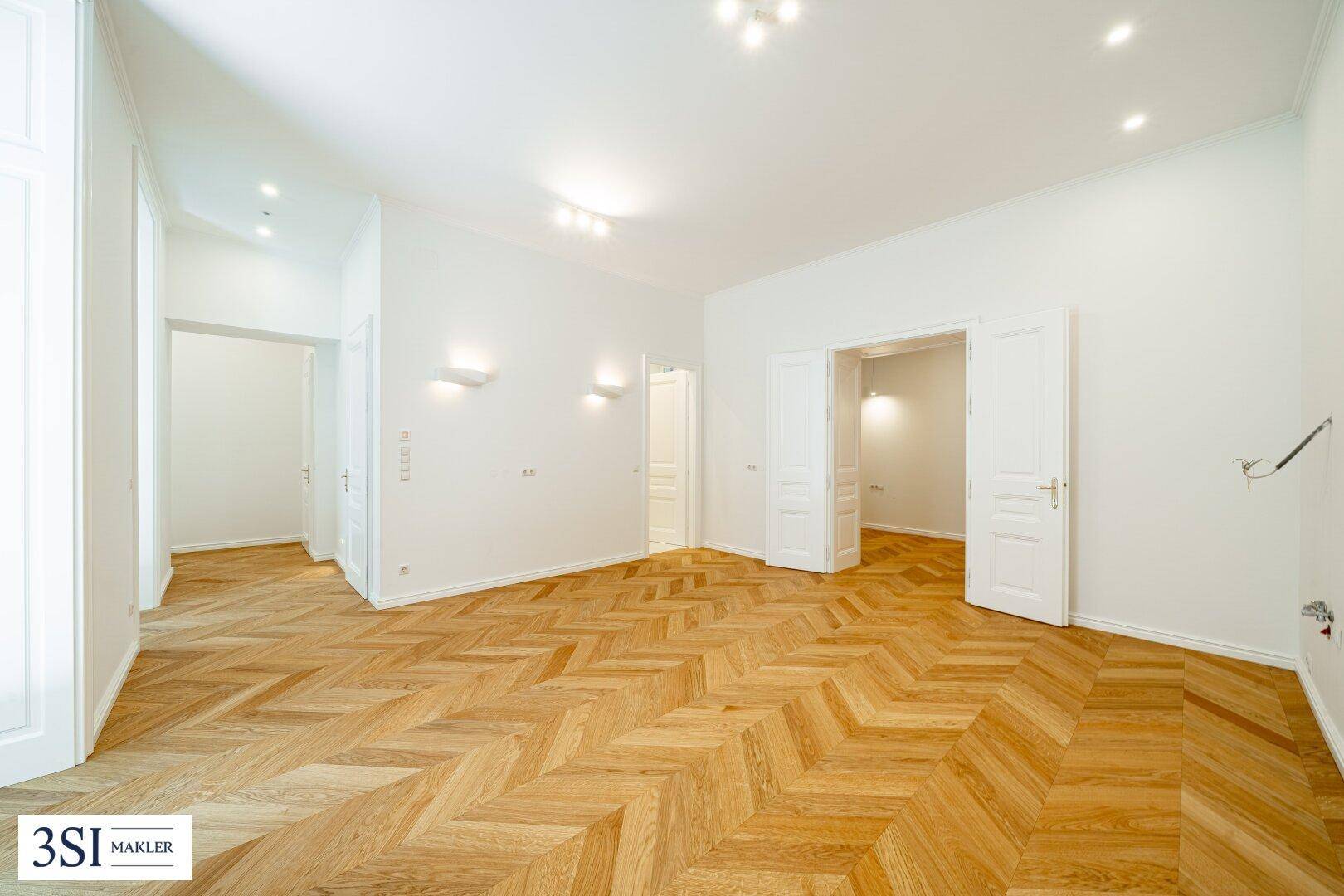 Geräumiges Zimmer mit elegantem Fischgrätparkett, weißen Wänden und mehreren Türen.