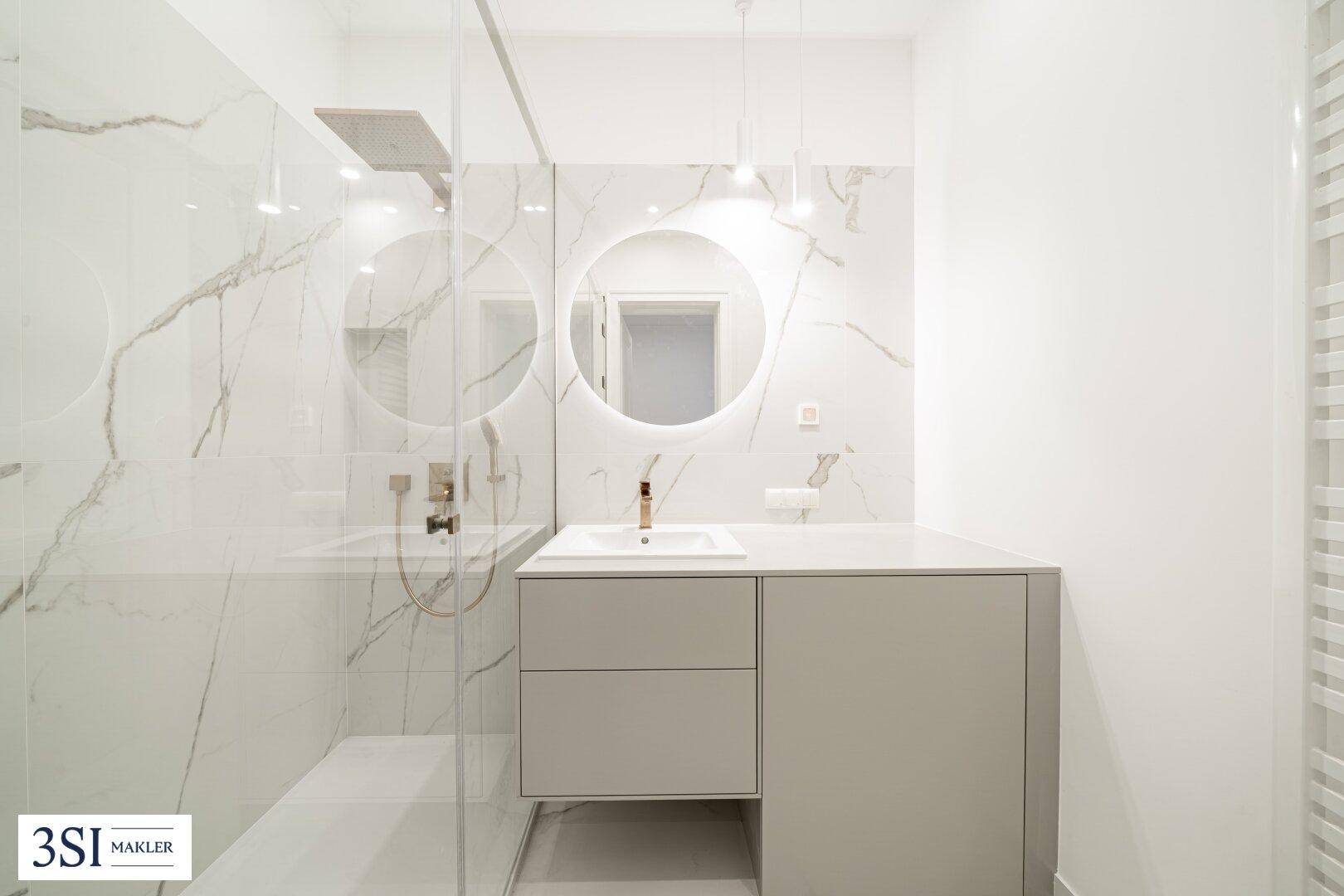 Modernes Badezimmer mit großer begehbarer Dusche, Marmorfliesen und rundem beleuchtetem Spiegel.